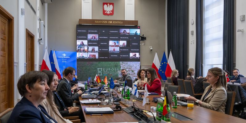 Udeleženci srečanja EUROSAI WGEA, ki poteka v sejni sobi vrhovne revizijske institucije Poljske (Najwyższa Izba Kontroli), sedijo za dolgo konferenčno mizo z zastavicami držav udeleženk, računalniki in dokumenti. Na zadnji steni se na zaslonu prikazujejo udeleženci, povezani na daljavo, poleg pa logotip srečanja in okoljski motiv.