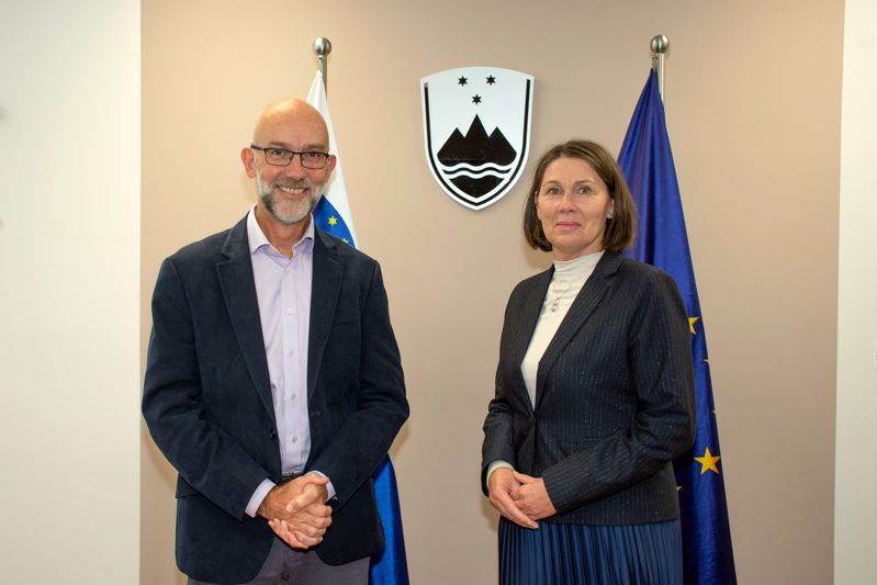 Skupinska fotografija generalnege revizorja za Wales Adriana Cromptona in predsednice računskega sodišča Jane Ahčin pred slovensko in evropsko zastavo ter slovenskim grbom