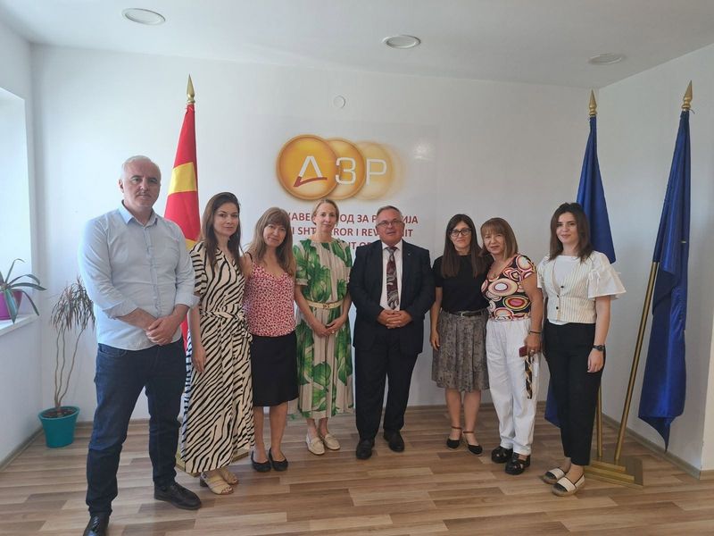Skupinska fotografija udeležencev delovnega srečanja v prostorih VRI Severne Makedonije v Skopju. Na fotografiji sta predstavnici Računskega sodišča Republike Slovenije skupaj z generalnim revizorjem VRI Severne Makedonije mag. Maksimom Acevskim ter člani revizijske ekipe. Udeleženci stojijo pred logotipom institucije, ob zastavi Severne Makedonije in zastavah Evropske unije.