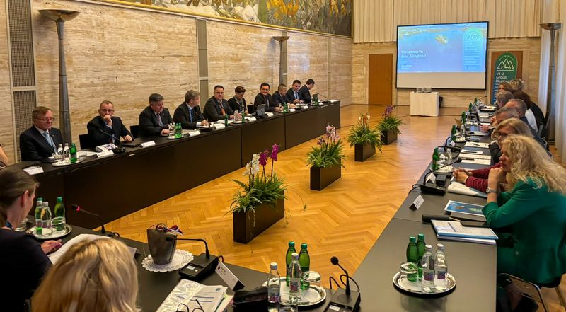 Delovni sestanek Delovni sestanek udeležencev srečanja V4+2 na Bledu. Delegati sedijo za dolgimi mizami, opremljenimi z mikrofoni, dokumenti in pijačo. V ospredju so cvetlični aranžmaji, na projekcijskem platnu pa uvodna predstavitev srečanja z napisom »Welcome to Bled, Slovenia!«.