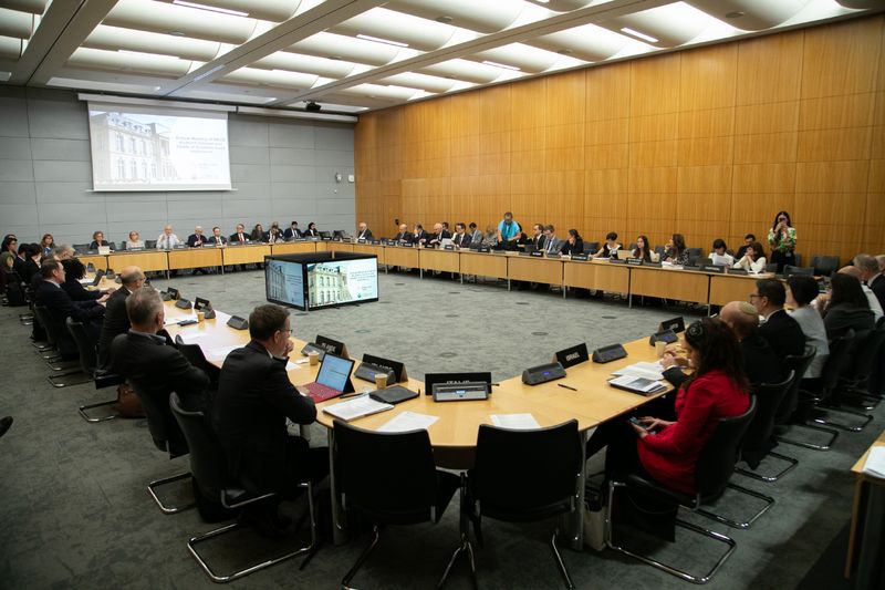 V veliki konferenčni dvorani na sedežu OECD v Parizu poteka mednarodno srečanje. Več deset udeležencev sedi okoli velike pravokotno razporejene konferenčne mize, ki obdaja osrednji prostor z zaslonom za predstavitve. Pred delegati so prenosni računalniki, dokumenti in označbe delegacij. Na sprednji steni je projicirana predstavitev, udeleženci pa pozorno spremljajo razpravo. Sodobna dvorana z lesenimi stenami in svetlo osvetlitvijo ustvarja formalno in profesionalno vzdušje mednarodnega srečanja.