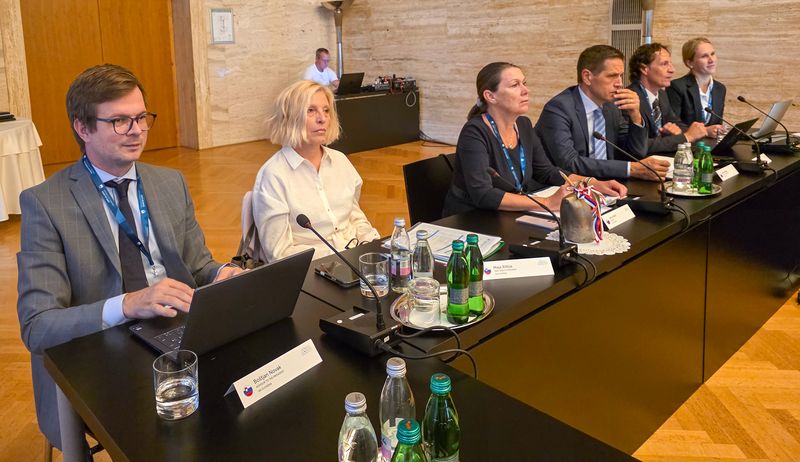Predsednica računskega sodišča Jana Ahčin z namestnikoma mag. Majo Bilbija in mag. Aleksejem Šinigojem ter člani delegacije Predsednica Računskega sodišča Republike Slovenije Jana Ahčin z namestnikoma mag. Majo Bilbija in mag. Aleksejem Šinigojem ter člani delegacije na delovnem sestanku V4+2. Udeleženci sedijo za konferenčno mizo z mikrofoni, prenosnimi računalniki, dokumenti in pijačo.