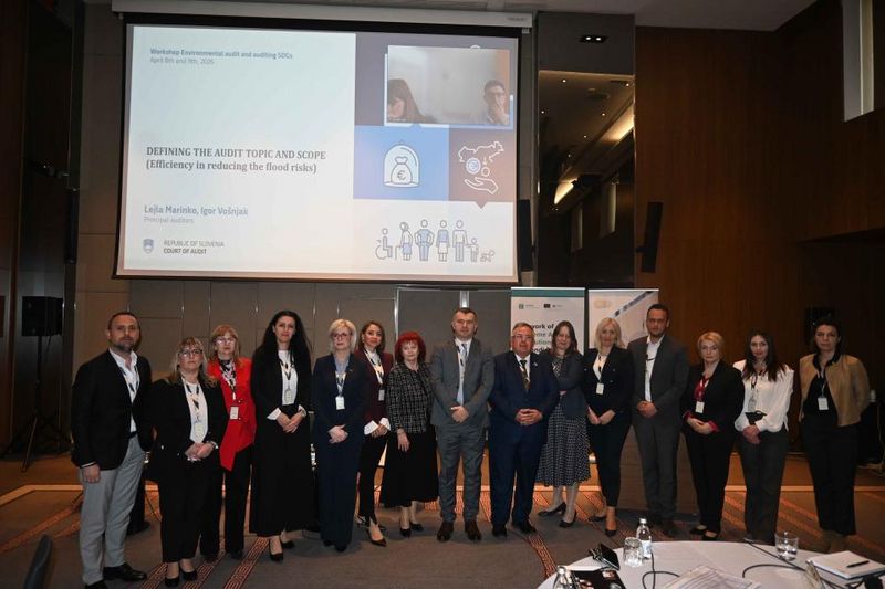 Skupinska fotografija udeležencev delavnice pred projekcijskim platnom s predstavitvijo o reviziji učinkovitosti zmanjševanja poplavnih tveganj.