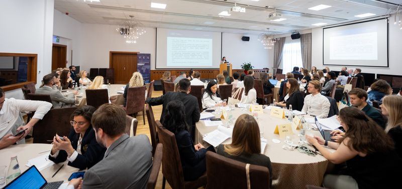 Pogled na konferenčno dvorano med predstavitvijo. Udeleženci sedijo za okroglimi mizami, spremljajo predavanje in razpravljajo o strokovnih temah. Na sprednjem delu dvorane govornica predstavlja vsebino ob projekciji na velikem platnu.