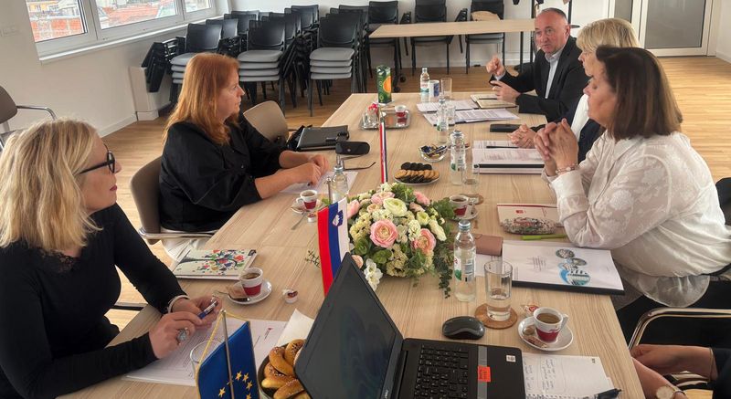 Delovni sestanek na sedežu VRI Hrvaške Skupina sedmih ljudi sedi okoli velike lesene konferenčne mize v svetlem sejni sobi. Na mizi so dokumenti, prenosni računalnik, skodelice s kavo, steklenice vode, krožniki s piškoti in cvetlični aranžma. Na sredini mize stojita majhni zastavici Slovenije in Evropske unije. V ozadju so ob steni zloženi stoli in velika okna. Ena oseba na koncu mize govori in gestikulira z roko, ostali poslušajo in sodelujejo v pogovoru.