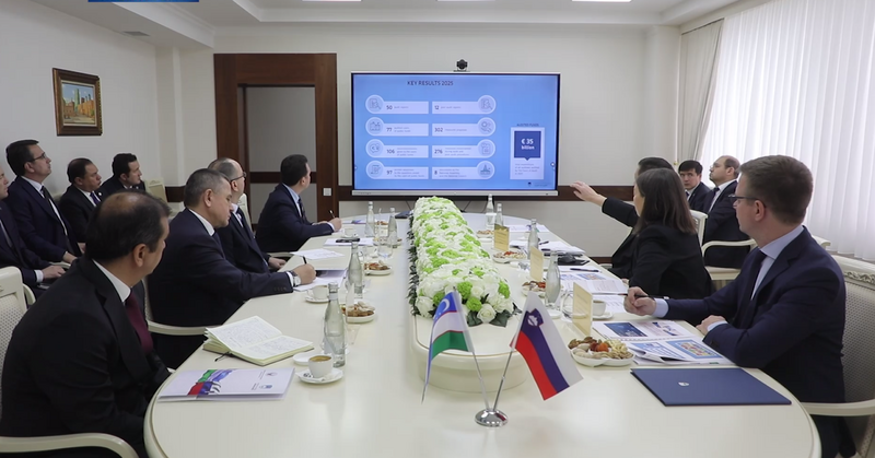 Udeleženci sedijo ob dolgi mizi in gledajo proti velikemu zaslonu na steni, kjer je prikazana predstavitev z naslovom »Key Results 2025«. Jana Ahčin sedi na desni strani in z roko kaže proti zaslonu. Na mizi so dokumenti, steklenice vode in majhni namizni zastavici Slovenije in Uzbekistana.