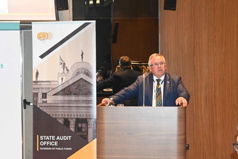 Generalni revizor VRI Severne Makedonije mag. Maksim Acevski za govorniškim pultom na konferenci, ob njem promocijski pano »State Audit Office – Guardian of Public Funds«.