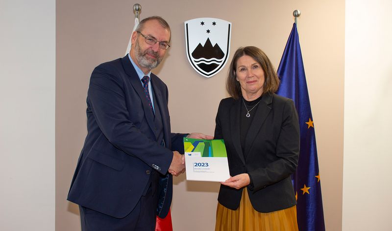 Member of the European Court of Auditors, Jorg Kristijan Petrovič, MA, presents the European Court of Auditors' Annual Reports for 2023 to the President of the Court of Audit of the Republic of Slovenia, Jana Ahčin. Both are standing in front of the Slovenian and European flags and the coat of arms of the Republic of Slovenia.