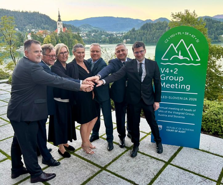 Predsedniki oziroma vodje delegacij sodelujočih VRI Skupinska fotografija predsednikov oziroma vodij delegacij sodelujočih VRI na srečanju V4+2 na Bledu. Stojijo na prostem in imajo roke položene druga na drugo v skupni gesti. V ozadju sta Blejsko jezero in Blejski otok s cerkvijo, ob skupini pa stoji pano srečanja V4+2 Group Meeting 2026.V4+2 Group Meeting 2026.