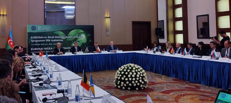 Udeleženci sedijo za mizo v obliki črke U na 22. letnem srečanju delovne skupine EUROSAI za okoljsko revidiranje v Bakuju, kjer razpravljajo o izzivih, povezanih z ekstremnimi vremenskimi dogodki. Za mizo so nameščene zastavice, ki označujejo sodelujoče države, medtem ko je ozadje okrašeno z logotipom EUROSAI in napisom dogodka v azerbajdžanščini in angleščini