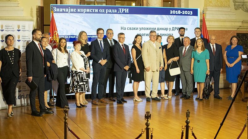 Group photo of representatives of foreign delegations and representatives of the Supreme Audit Institution of Serbia in the premises of the National Assembly, as part of the exhibition opening, in front of the flags and a backdrop displaying key data about the anniversary of the Supreme Audit Institution of Serbia.