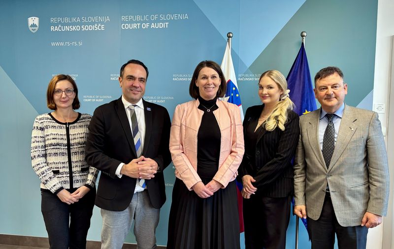 Skupinska fotografija predsednice računskega sodišča Republike Slovenije Jane Ahčin z delegacijo odbora za spremljanje, ki vključuje Ullo Santti, Konstantinosa Koukasa, profesorja Angela Morena in Svitlano Pereverten. Fotografija je posneta pred uradnim ozadjem s slovenskimi in evropskimi simboli.