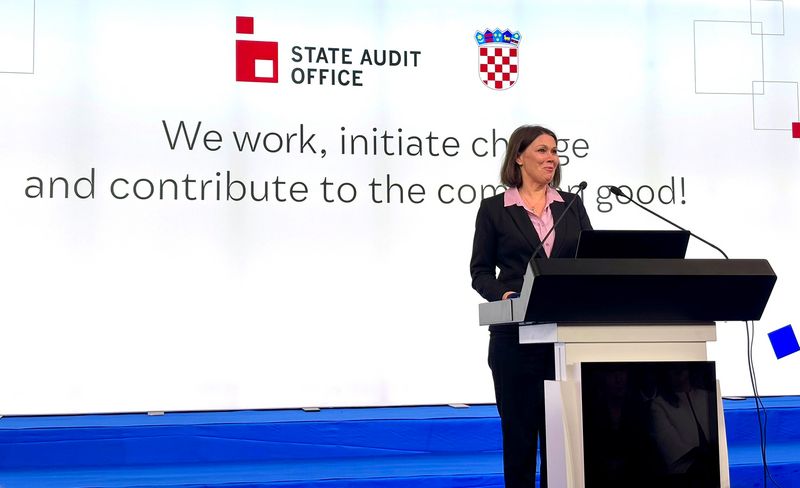 Fotografija predsednice Računskega sodišča Republike Slovenije, Jane Ahčin, med njenim govorom na jubilejni konferenci ob 30. obletnici vrhovne revizijske institucije Hrvaške. V ozadju je projekcija z napisom: "We work, initiate change and contribute to the common good!" ter logotipom VRI Hrvaške in državnim grbom Hrvaške.