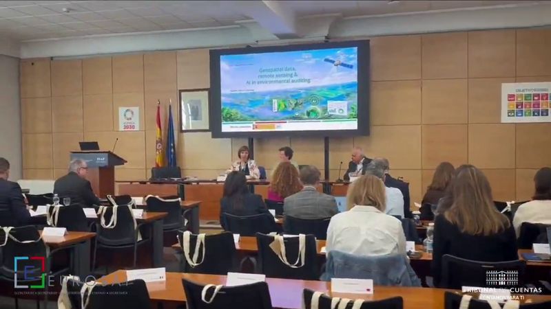 Fotografija prikazuje udeležence srečanja v konferenčni dvorani v Madridu. V ospredju sedijo udeleženci za mizami, obrnjeni proti govornikom in velikemu projekcijskemu zaslonu. Na zaslonu je predstavitev o geoprostorskih podatkih, daljinskem zaznavanju in umetni inteligenci v okoljskem revidiranju. Pred zaslonom sedijo trije govorci, ob strani prostora pa sta vidni španska in evropska zastava.