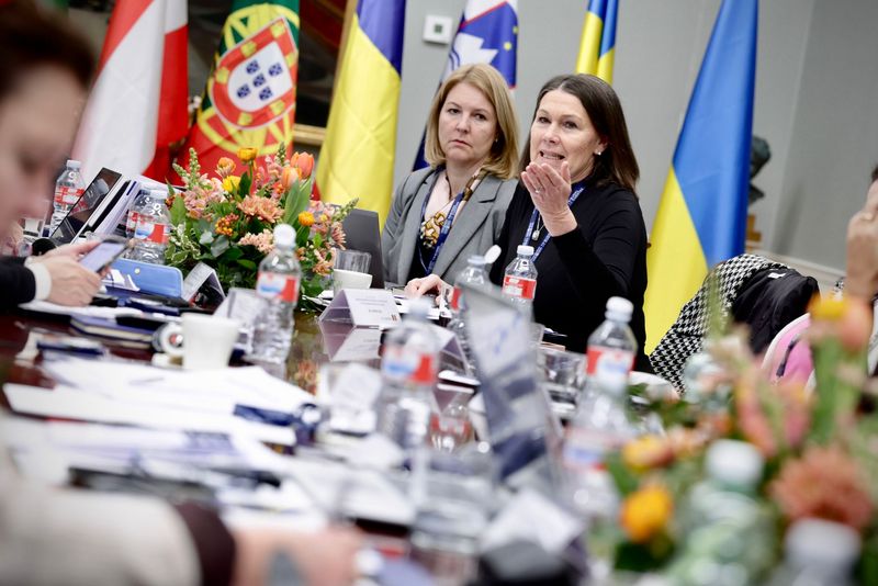 Predsednica računskega sodišča Jana Ahčin med razpravo na delovnem srečanju aktivno sodeluje in z gestikulacijo poudarja svoje stališče, ob njej pa druge udeleženke pozorno spremljajo razpravo. V ozadju so vidne zastave sodelujočih držav, ki poudarjajo mednarodni značaj dogodka.