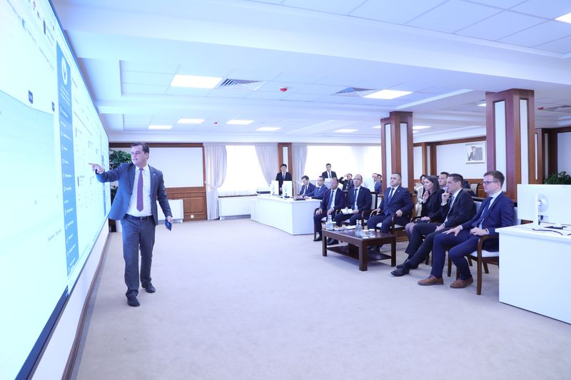 Center za oddaljeni nadzor VRI Uzbekistana; moški stoji ob velikem zaslonu in kaže na prikazane podatke, medtem ko skupina udeležencev sedi nasproti in spremlja predstavitev.