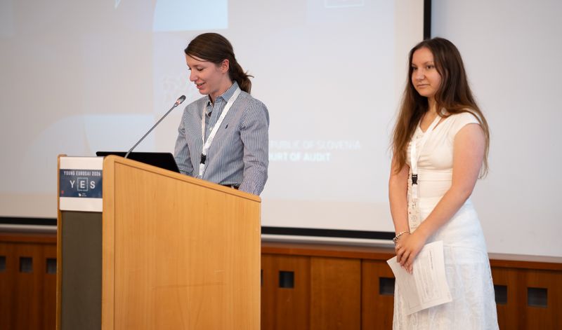 Polona Mlakar in Karmen Cej med predstavitvijo računskega sodišča V konferenčni dvorani dve mladi revizorki Računskega sodišča Republike Slovenije predstavljata prispevek na konferenci Young EUROSAI. Ena stoji za govorniškim pultom in govori v mikrofon, druga stoji ob njej z dokumenti v rokah. V ozadju je projekcijsko platno s predstavitvijo.