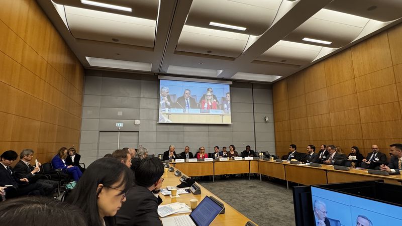 Sejna soba na sedežu OECD v Parizu z velikim ovalnim omizjem, okoli katerega sedijo udeleženci, na steni pa je projekcijsko platno s prenosom razprave.