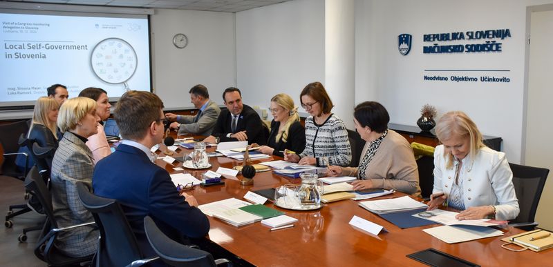 Fotografija prikazuje udeležence delovnega sestanka na računskem sodišču Republike Slovenije. Udeleženci sedijo za dolgo mizo z dokumenti in gradivi, v ozadju je prikazan zaslon s predstavitvijo o lokalni samoupravi v Sloveniji.