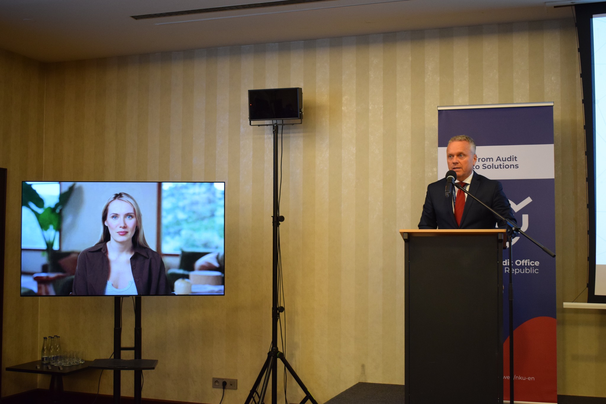 Ľubomír Andrassy med nagovorom udeležencev konference stoji za govorniškim pultom in govori v mikrofon. Ob njem je zaslon z govorko, generirano s pomočjo umetne inteligence, v ozadju pa stoji promocijski pano organizatorja dogodka.