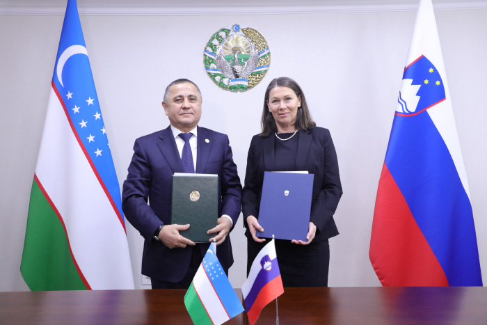 Predsedujoči vrhovni revizijski instituciji Uzbekistana dr. Mehriddin Abdullajev in predsednica Računskega sodišča Republike Slovenije Jana Ahčin stojita drug ob drugem in držita podpisana dokumenta. Za njima sta zastavi Uzbekistana in Slovenije ter državni grb na steni, pred njima na mizi pa stojita manjši zastavici obeh držav.