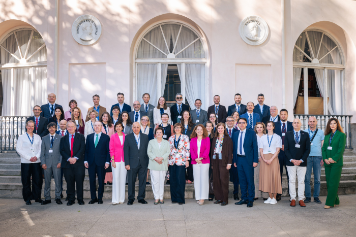 Skupinska fotografija udeležencev srečanja v Madridu pred zgodovinsko stavbo z velikimi obokanimi okni. Na stopnicah stoji približno petdeset udeležencev iz različnih držav, oblečenih večinoma v poslovna oblačila in z identifikacijskimi trakovi okoli vratu. Udeleženci so razporejeni v več vrstah in gledajo proti fotografu. Fotografija je posneta na prostem v sončnem vremenu, na pročelju stavbe pa se vidijo dekorativni reliefi in sence dreves.